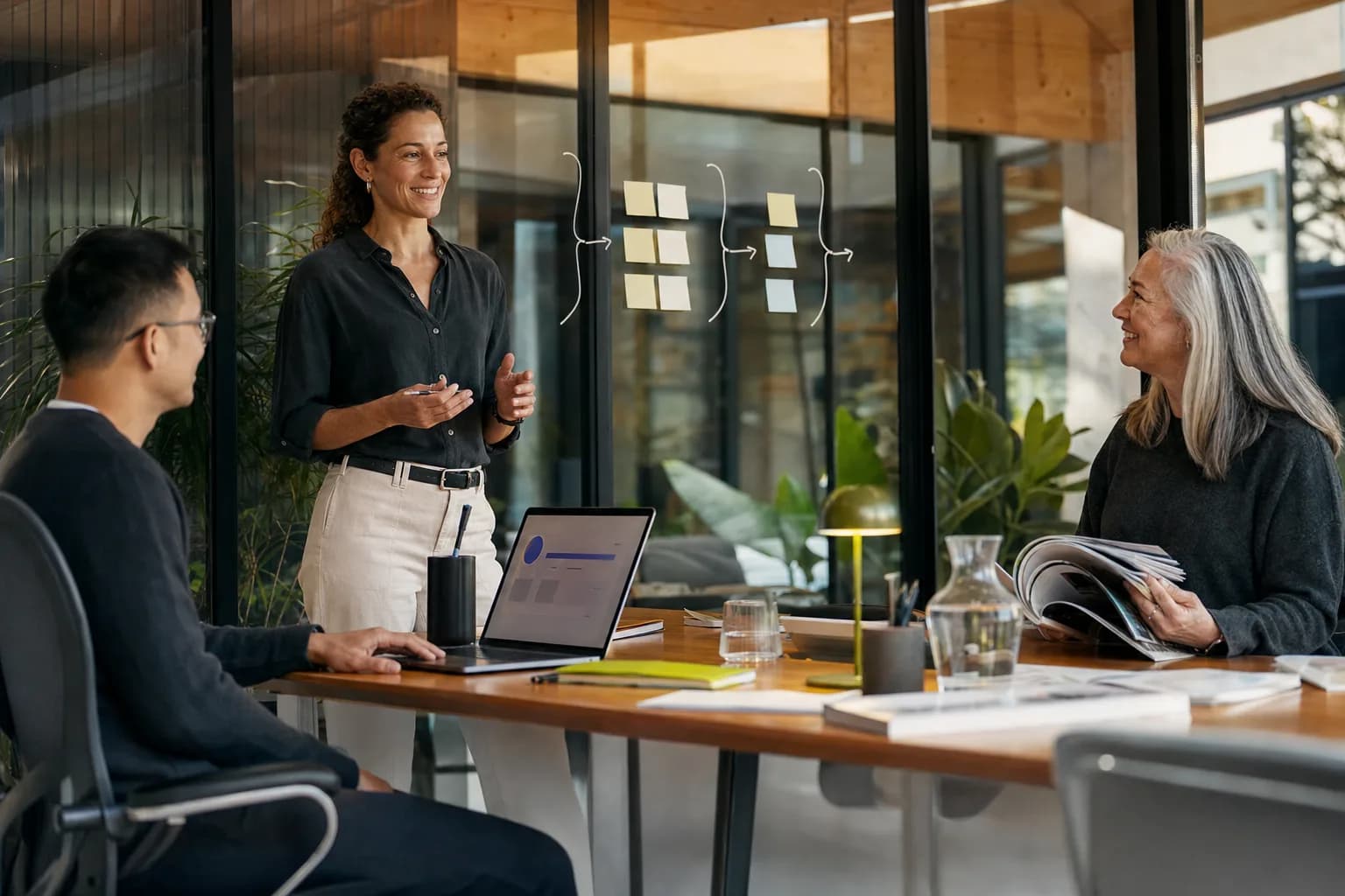 A founder walks two teammates through a three-layer plan at a sunlit office table, one on a laptop and one reading printed magazines: the systems, search and press behind personal brand infrastructure.