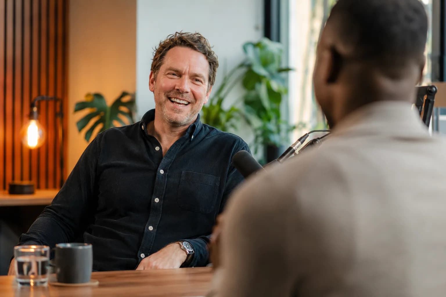 A founder in warm conversation with a podcast host in a bright modern studio, illustrating how a borrowed audience puts a founder in front of buyers who already trust the host.